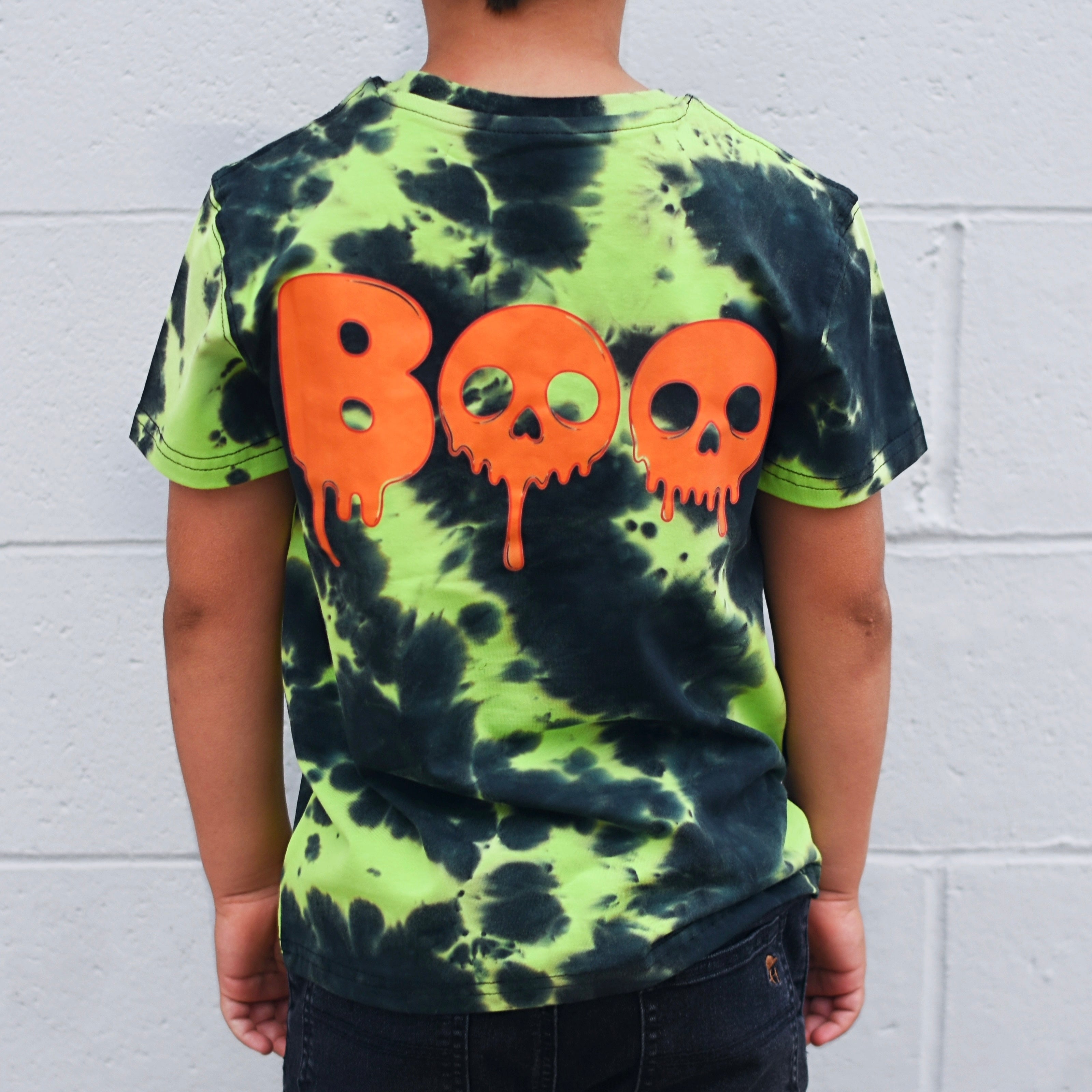 A child wearing a neon green and black tie-dye t-shirt from the back, featuring the word 'BOO' in dripping orange letters with skull details inside the letters, set against a white brick wall.