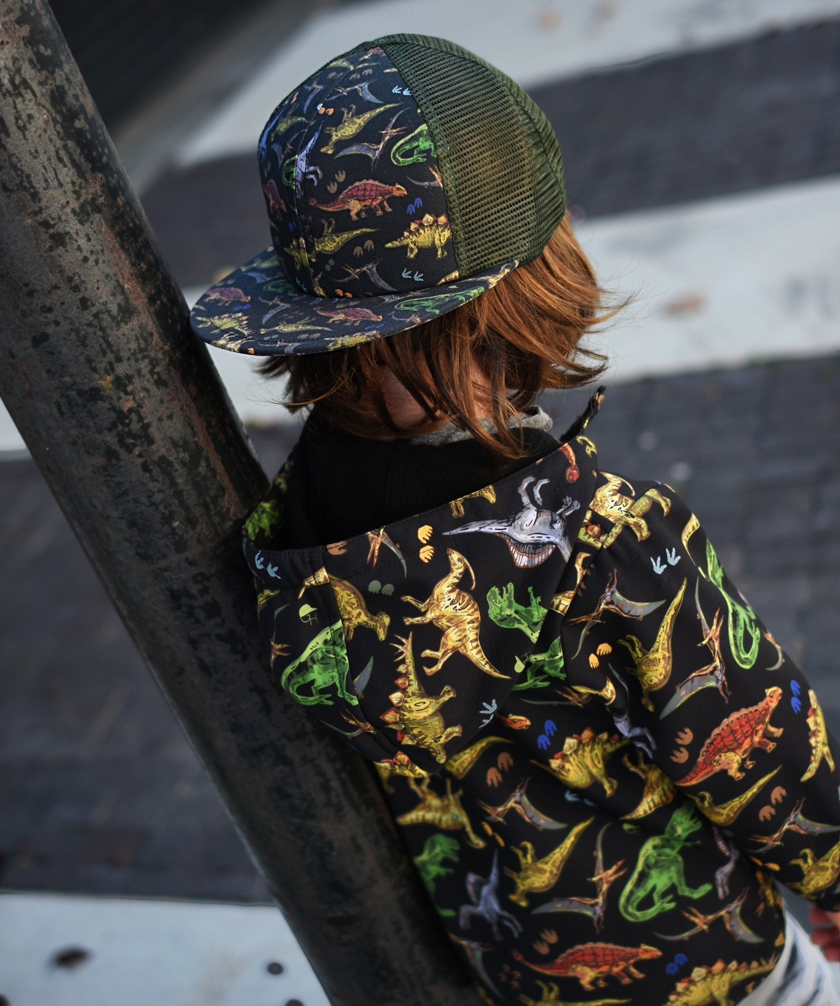 image of a boy leaning against a metal pole wearing the Dinosaur Jacket - George Hats little boys winter coat