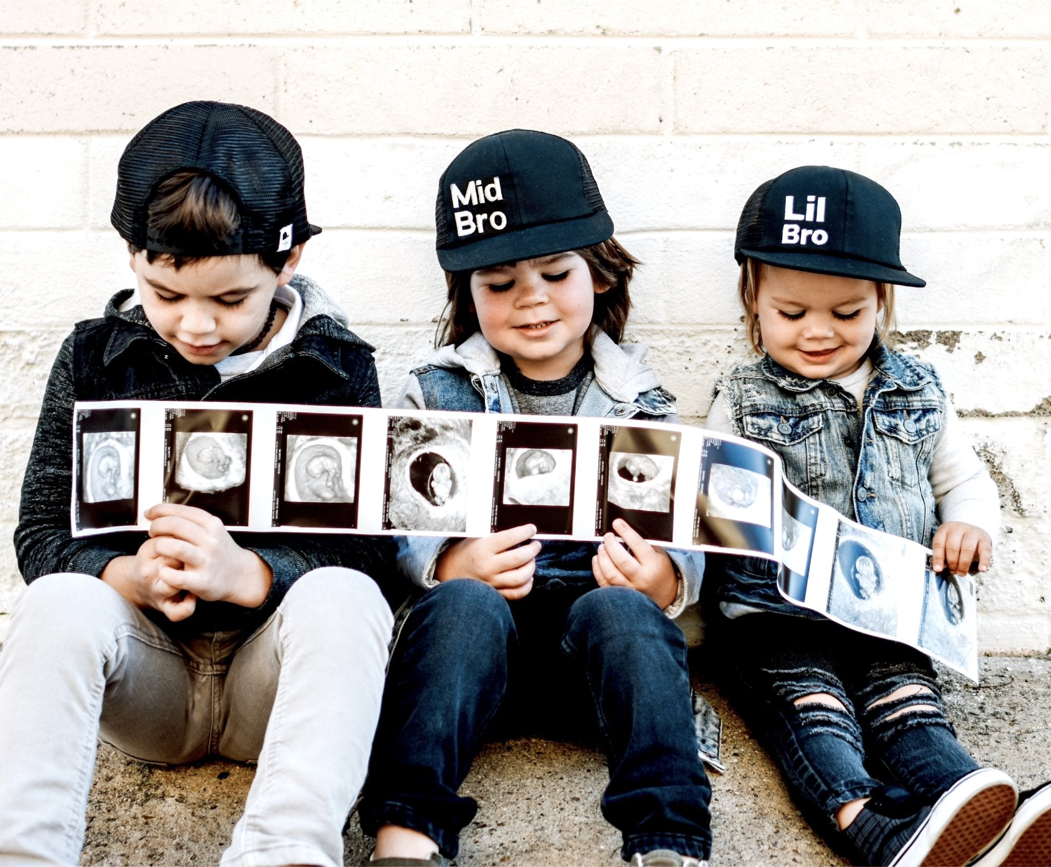 Picture of three boys in lil bro trucker, mid bro trucker, and big boy trucker hats holding an ultrasound image