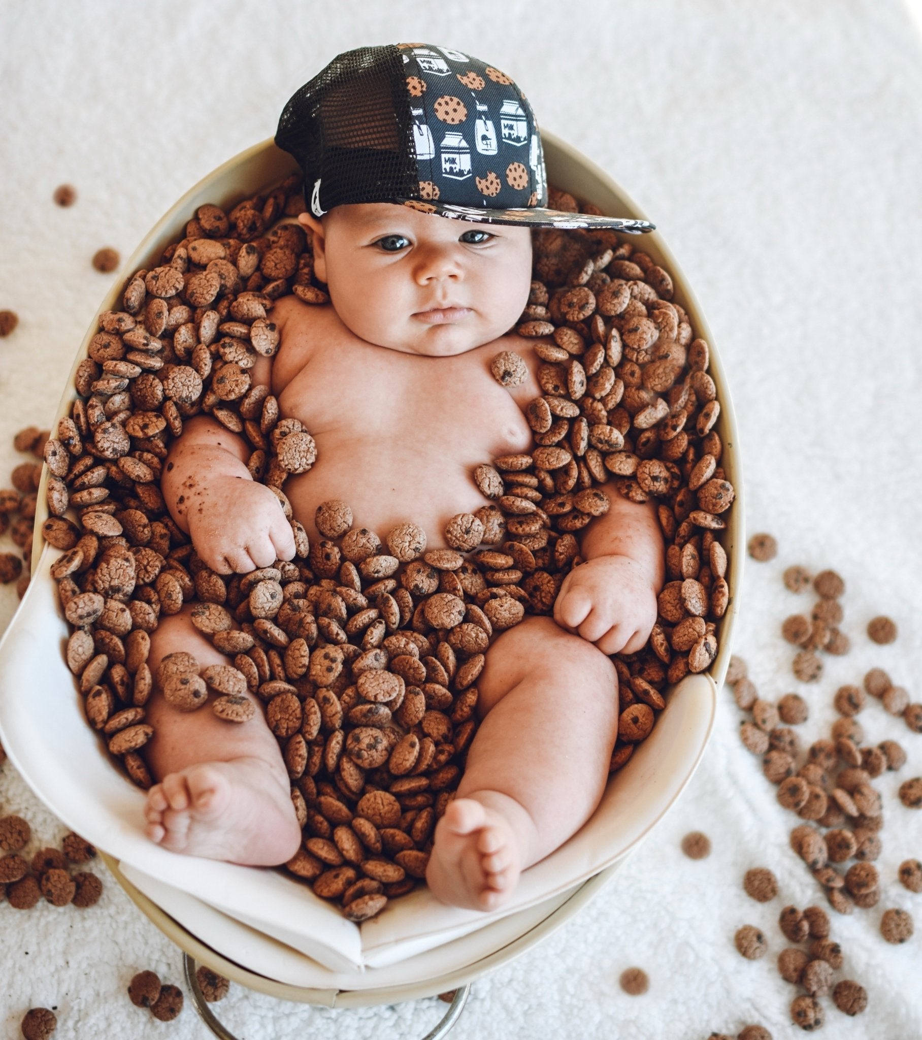 Image of a baby laying in a bowl filled with small cookies and wearing the Milk and Cookies kids trucker hat from George Hats