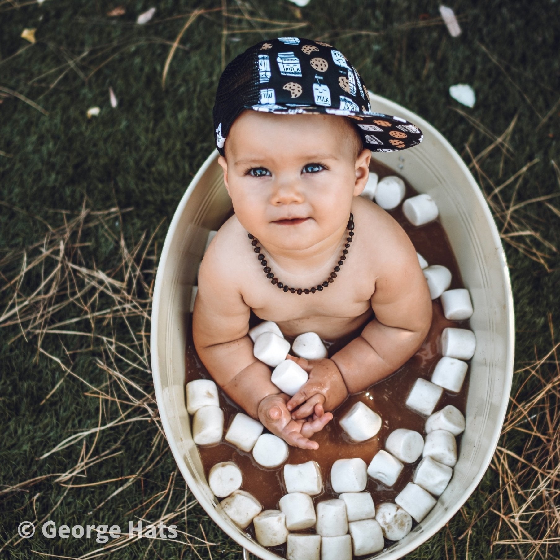 Image of a baby wearing the Milk and Cookies kids trucker hat sitting in a small metal pool filled with hot chocolate and marshmallows 