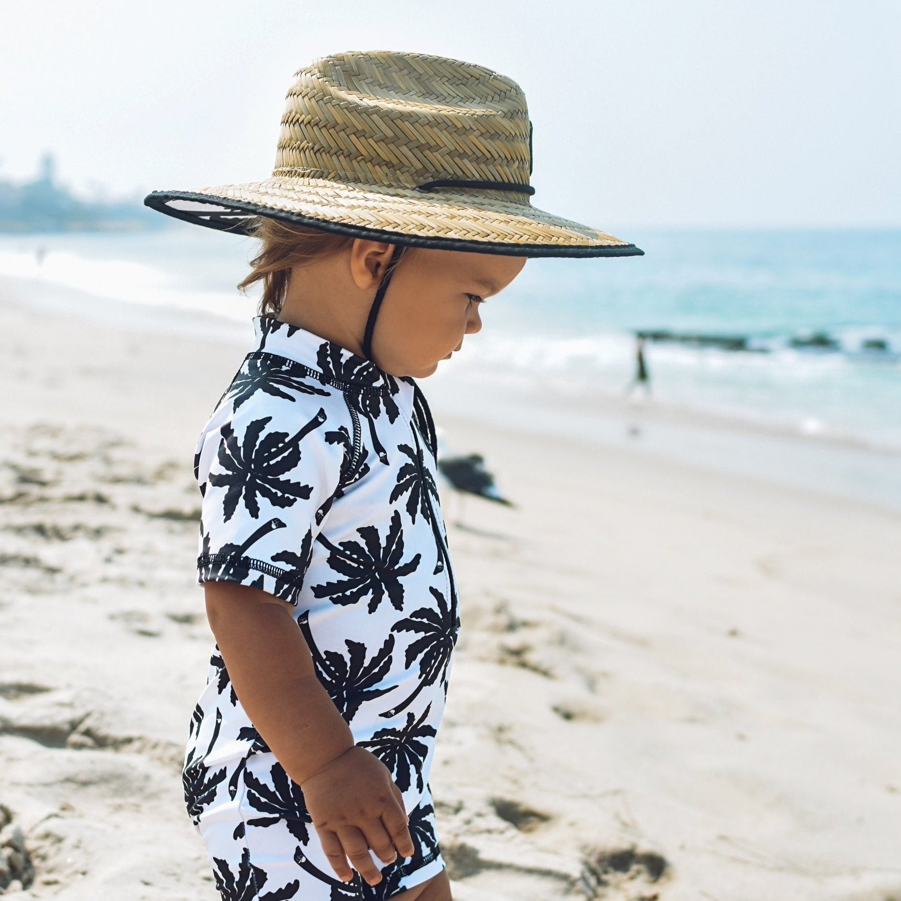 An image of a child in the George Hats Palm Straw Hat and Sunsuit on the beach.