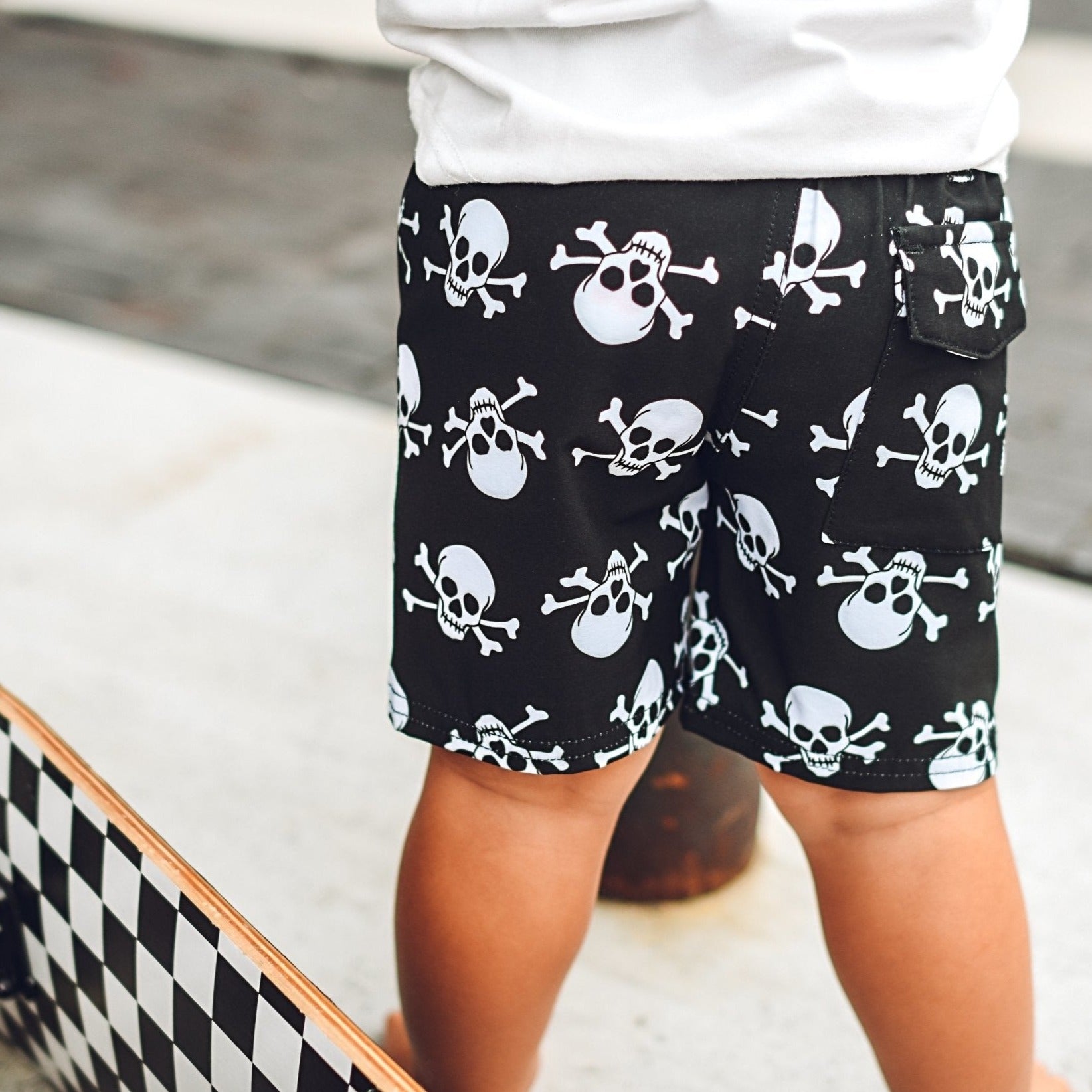 Boy wearing black boy's shorts with white skull print beside a skateboard -Skull Walk Shorts - George Hats